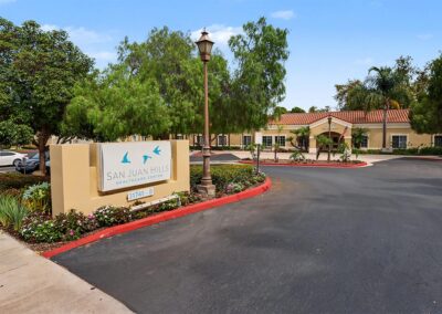 Entrance sign for San Juan Hills Healthcare Center at 31741-B with flower beds, a lamppost, and the curved drive to the tile-roof building