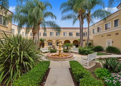 Landscaped courtyard with a tiered fountain, tall palm trees, and white benches framed by two-story Mediterranean-style buildings