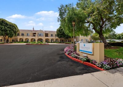 Circular drive entrance to Capistrano Senior Living with the monument sign, flower-lined curb, and two-story stucco building