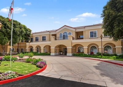 Arched front entry of a two-story Mediterranean-style senior living building with a second-floor balcony and red tile roof