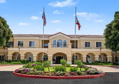 Senior living building front with American and California flags above a landscaped circular garden bed in the drive