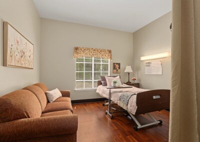Private resident room with an adjustable care bed, nightstand, window with blinds, floral wall art, and a sofa for visitors