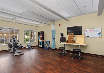 Rehabilitation therapy gym with a recumbent stepper, exercise bike, hand therapy boards, weights rack, and courtyard-view windows
