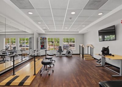 Bright physical therapy gym with parallel walking bars, practice stairs, a recumbent stepper, and large windows