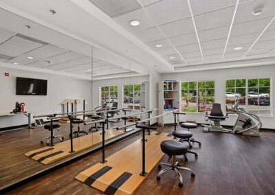 Parallel bars reflected in a wall mirror in a rehab gym with therapy stools, a recumbent stepper, and storage cabinets