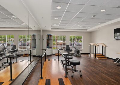 Rehab gym with parallel walking bars beside a mirrored wall, therapy stools, practice stairs, and supply cabinets