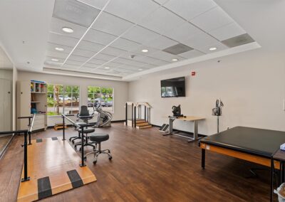 Physical therapy room with a treatment table, parallel bars, practice stairs, vital signs monitor, and wall-mounted TV
