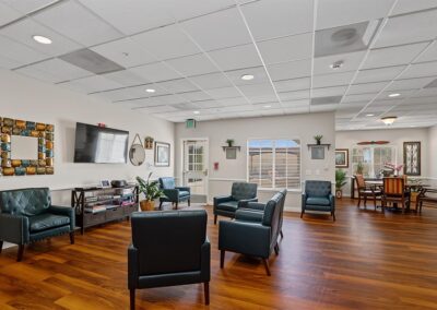 Spacious resident lounge with teal leather armchairs, a TV and media shelf, and a dining area on wood-look floors