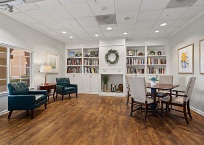 Resident library with built-in white bookshelves, teal leather armchairs, and a wood table with upholstered chairs