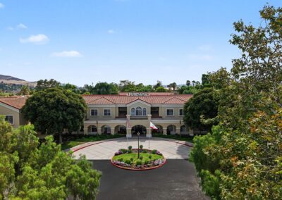 Aerial view of the senior living campus entrance with a circular drive, flagpoles, garden island, and tile-roof building among mature trees